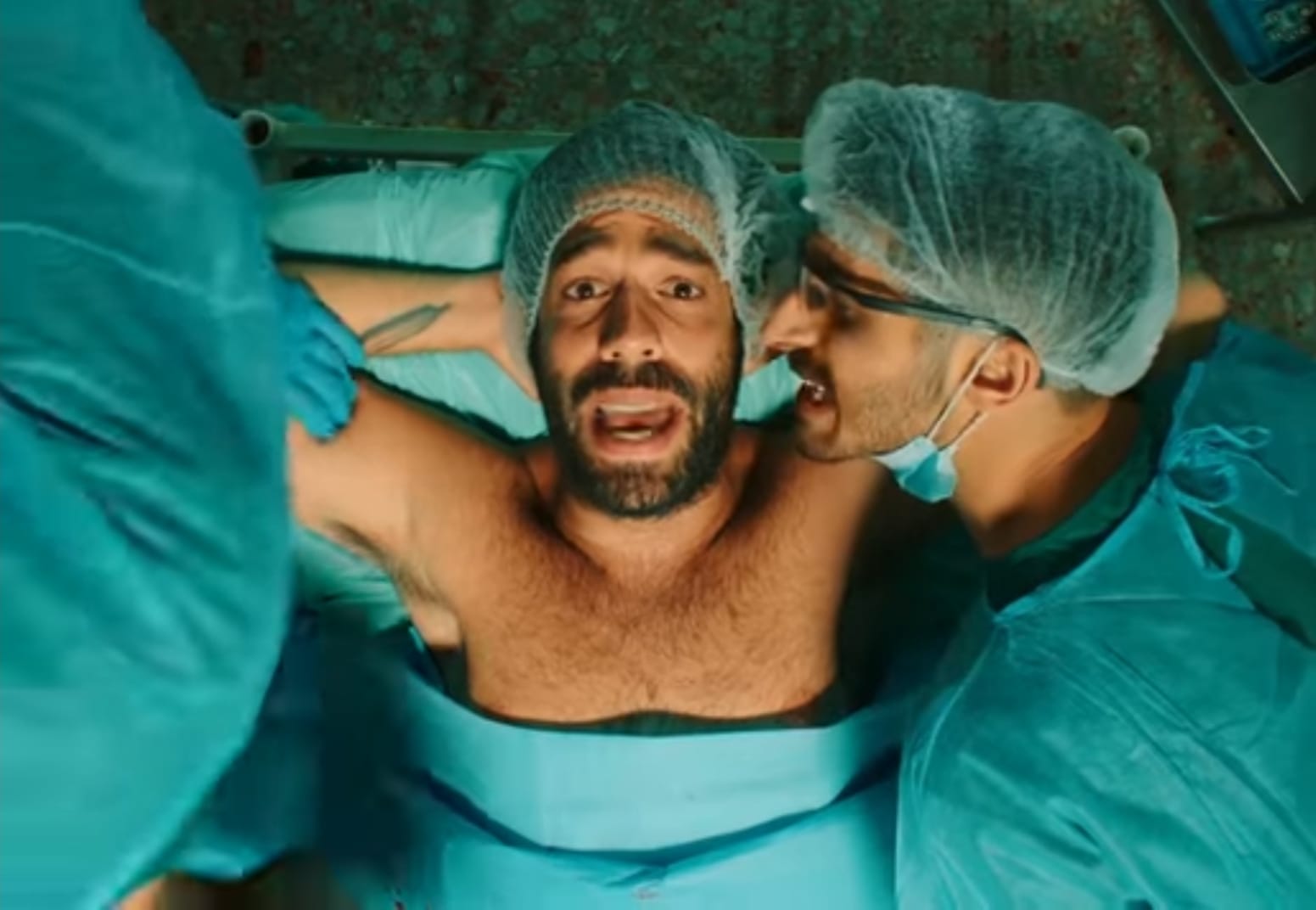 הזמר רביד פלוטניק שוכב על מיטת ניתוחים בחזה חשוף ידיו מאחורי ראשו. ולצידו שחקן בוחן אותו