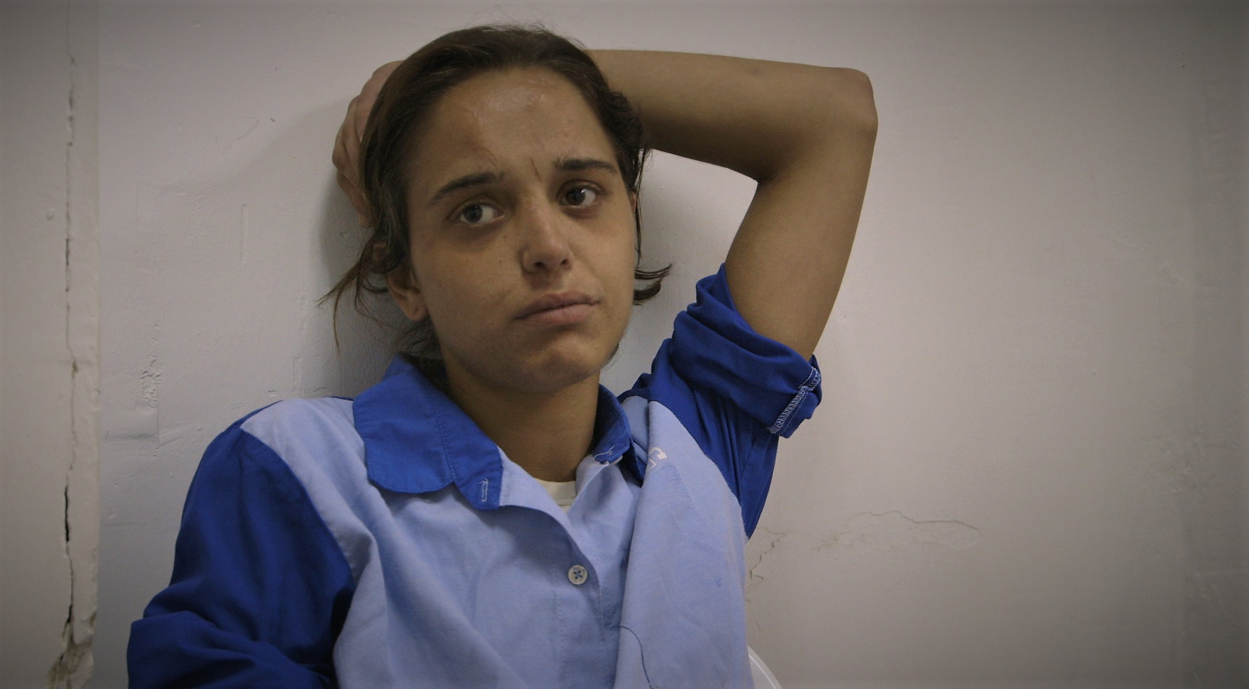 נערה יושבת במדי אסירה כחולים, משעינה ראש על ידה כנגד הקיר