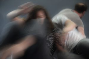 שלושה גופים נפגשים למגע דרך הגב במין קריסה לישיבה על רקע קיר אפור במרכז "שדרות אדמה"