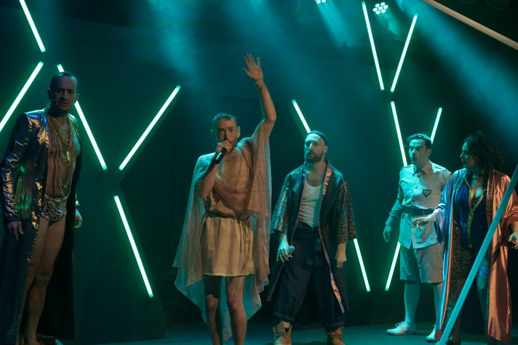 שחקן מחזיק מקרופון בידו מרים את היד, ארבעה שחקנים מסביבו מסתכלים עליו, פסי ניאון ירוקים מאירים את הבמה
