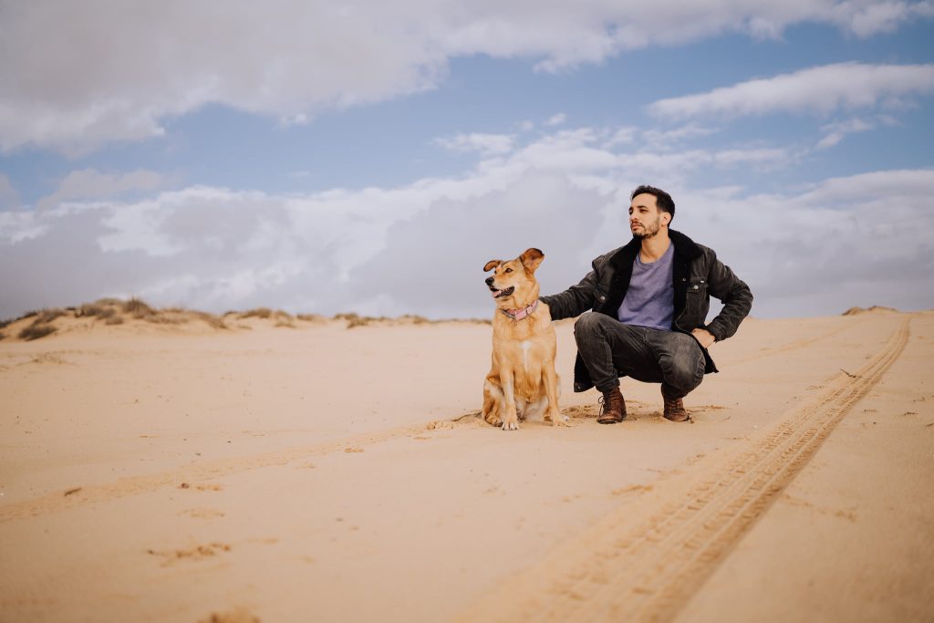 אדם כורע נמוך ולידו כלב יושב, שניהם מביטים לאותו כיוון, במדבר ומסביבם אדמה וחול, וברקע שמיים כחולים ועננים