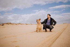 אדם כורע נמוך ולידו כלב יושב, שניהם מביטים לאותו כיוון, במדבר ומסביבם אדמה וחול, וברקע שמיים כחולים ועננים