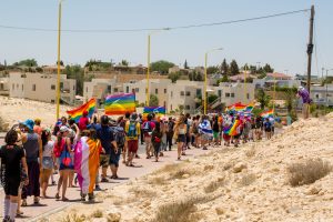 מצעד הגאווה במצפה רמון 2021. אנשים הולכים עם עשרות דגלים צבעוניים על כביש במדבר על רקע הבתים של העיירה