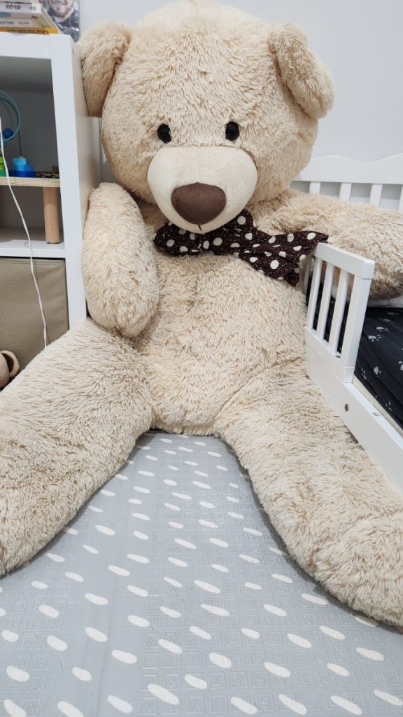 בובה פרוותית ענקית של דובי עם פפיון על מיטת ילדים