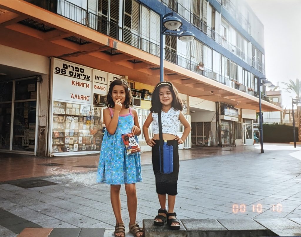 שתי אחיות עומדות ומחייכות אל המצלמה, באמצע הכיכר. הילדה הימנית נושאת מימייה כחולה והאחות השנייה מחזיקה שקית ממתקים.